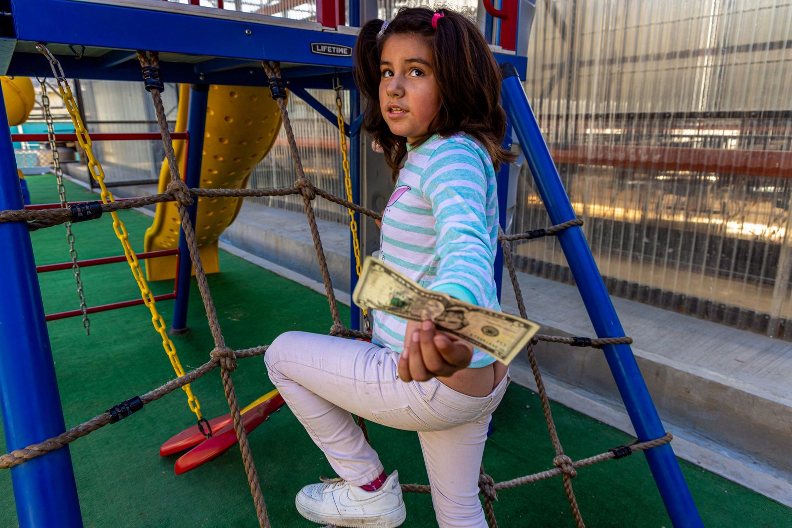 A young girl climbs the rope netting of a playground climbing frame, holding a five-dollar bill out toward the camera — The Liminal Innocence