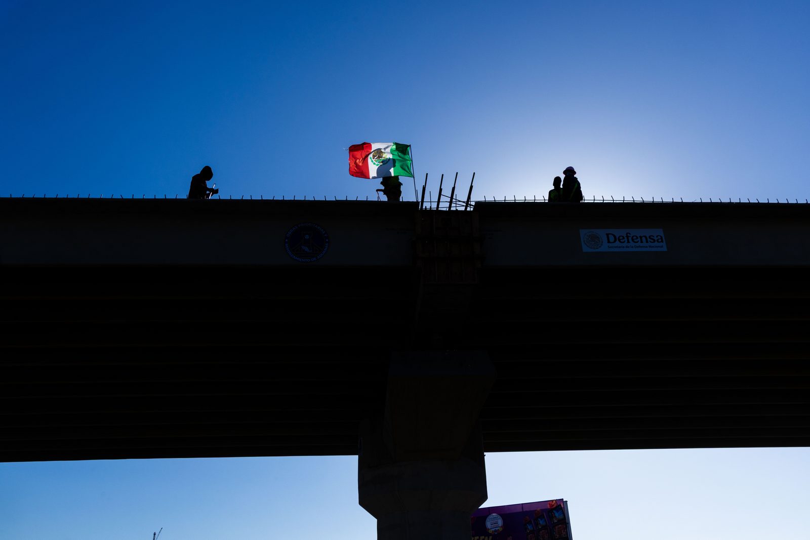 Workers silhouetted on an unfinished elevated highway against a clear blue sky, a Mexican flag flying above them — The Liminal Innocence