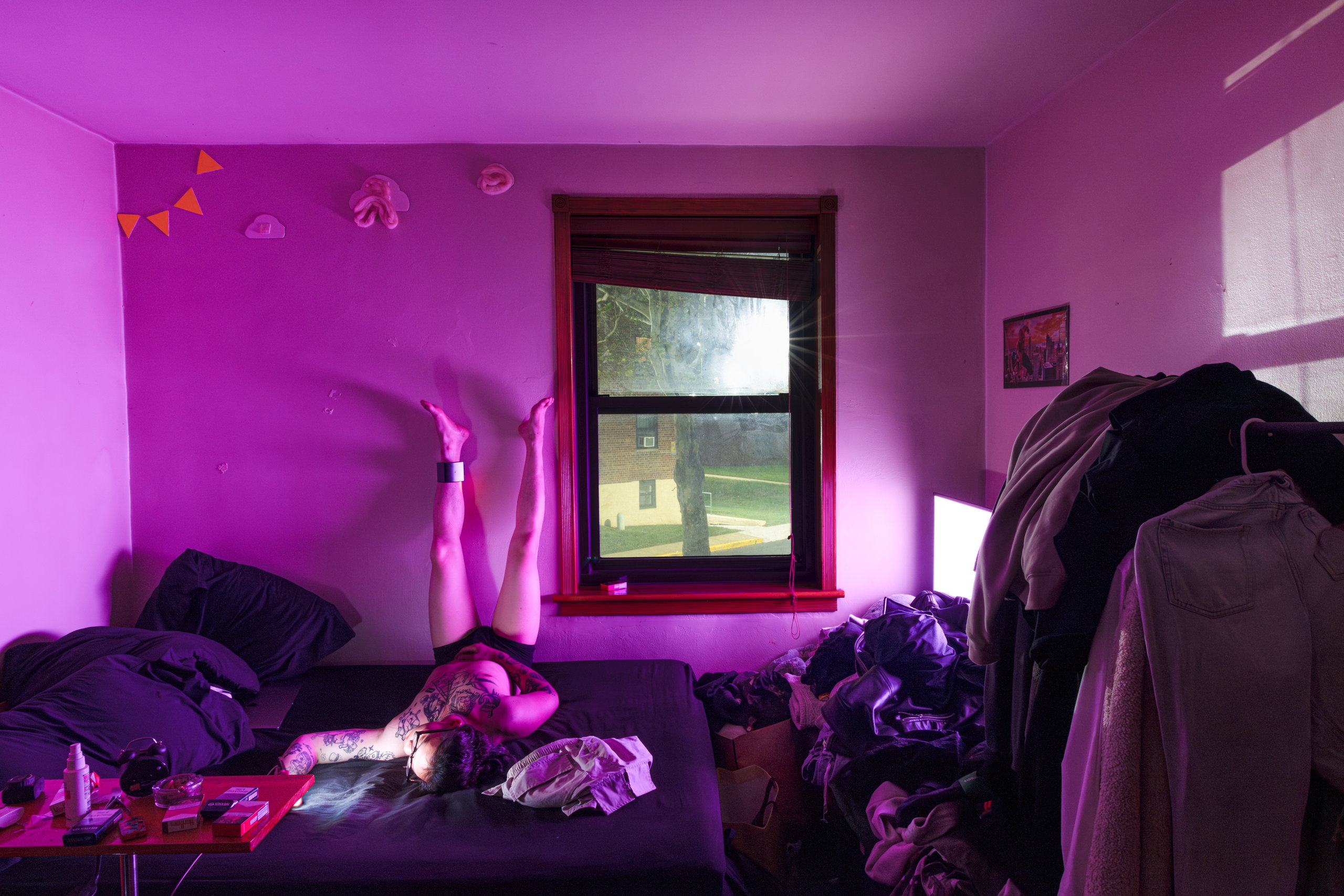 A bedroom washed in magenta light; a tattooed man lies on the bed with his legs up against the wall, sunlight bursting through the window — Win or Lose